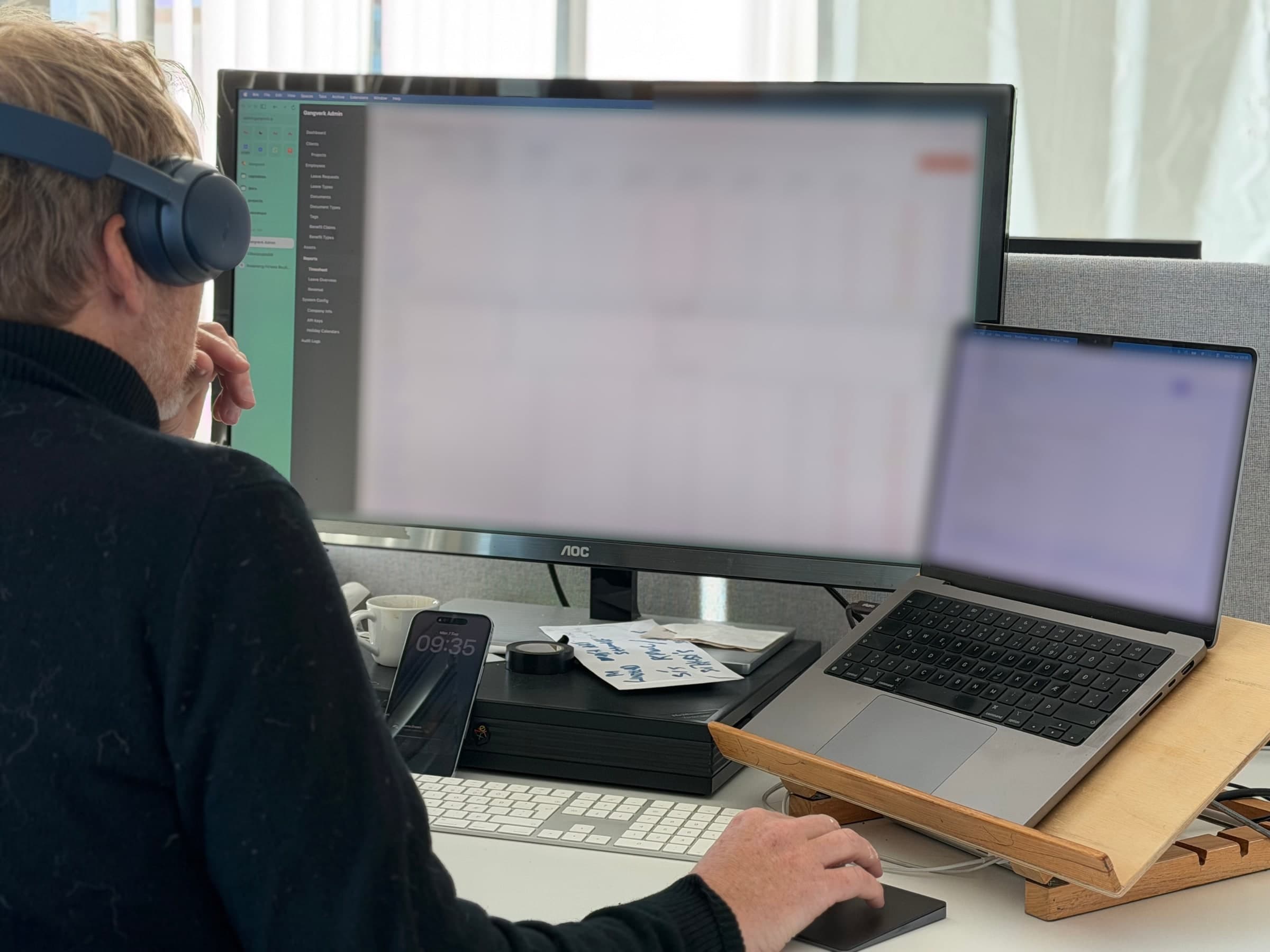 Atli at his desk in headphones, looking at a monitor showing the Gangverk Admin timesheet completeness table, with time tracking open on the laptop beside it